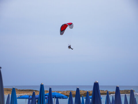Powered motor sailwing gliding in blue sky above blue mediterranean sea with nissie beach and blue umbrellas.