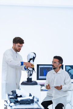 Next-generation prosthetics development, a lab-coated specialist opens the mechanical fingers of a white robot hand while a colleague dictates the measured values