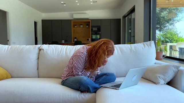 Smiling red-haired woman enjoying a cheerful video call on her laptop, relaxing comfortably on a modern white sofa in a bright, inviting home living room during the day.