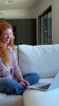 A young woman with vibrant red hair is genuinely laughing and clapping joyfully during an engaging video call on her laptop from the comfort of her modern home.