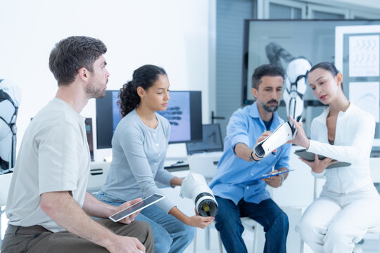 Bio-robotics laboratory: two junior researchers compare notes while the project lead raises the bionic forearm behind them so the whole group can see the shoulder collar