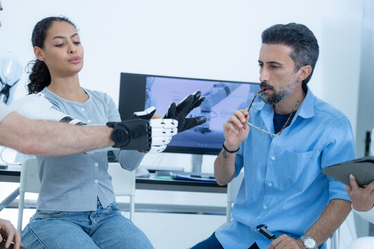 Bio-robotics laboratory: a black and white robotic hand is stretched out to its full reach while the lead engineer studies the open palm from the opposite chair
