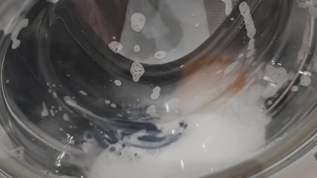 Close up of clothes spinning inside a washing machine drum with soapy water. Household chore and hygiene concept showing the cleaning cycle