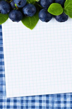 Fresh blueberries with water droplets and green leaves arranged above a blank white graph paper sheet on a blue checkered tablecloth