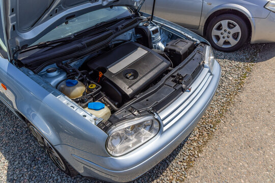 Close-up view of silver used German car with hood open, showcasing engine compartment. Bright sunlight highlights engine details, parked on gravel and asphalt.