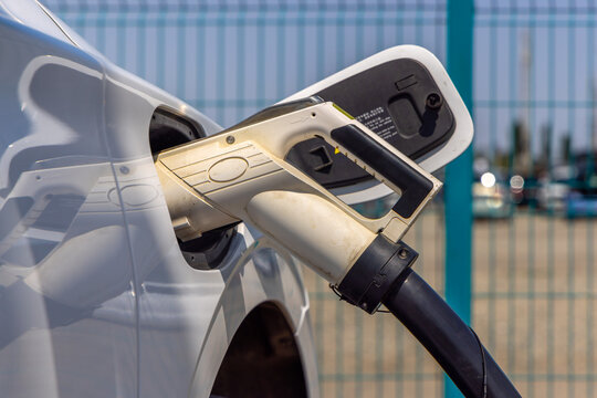 Close-up view of a white electric vehicle charging connector plugged into the car's port. Bright sunlight reflects off the car's surface, highlighting the charging process outdoors.