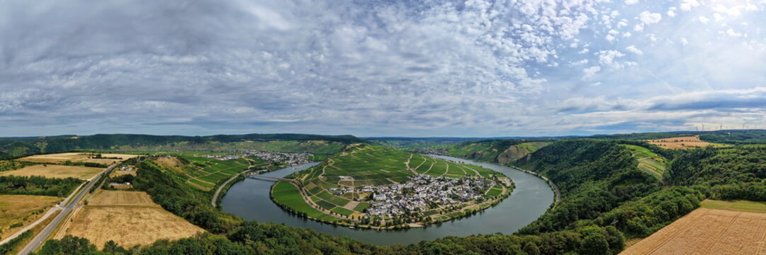 Panoramablick auf die Moselschleife bei Minheim mit idyllischen Weinbergen, blauem Flusslauf und typischem Weindorf unter leicht bew&ouml;lktem Sommerhimmel.