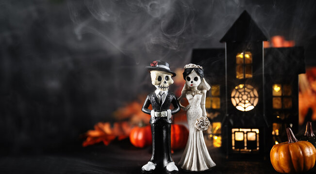 Skeleton bride and groom figurines beside glowing haunted house, pumpkins and smoke. Gothic Halloween wedding decorations on dark background with ample copy space. Halloween holiday card.