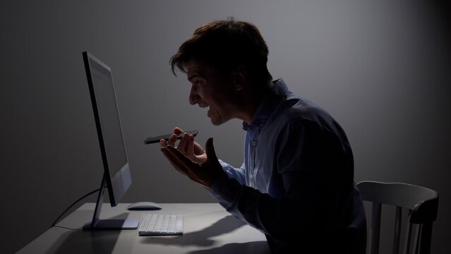Young man experiencing anger and stress, yelling into a smartphone while working on a computer in a dimly lit office, symbolizing professional frustration and communication problems