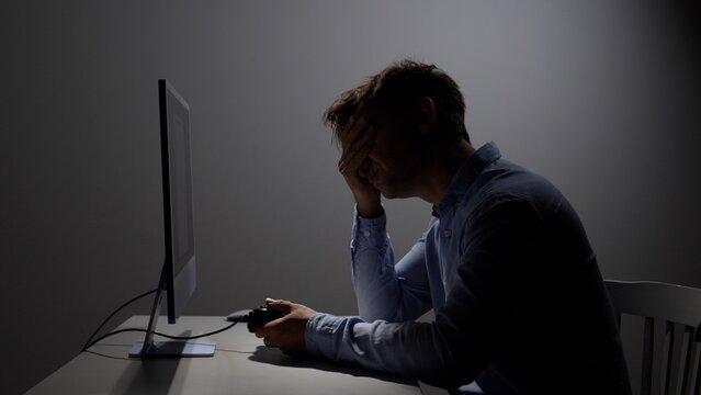 Young man sitting in dark room experiencing frustration and disappointment while playing video game with a controller, covering his face with his hand near computer monitor