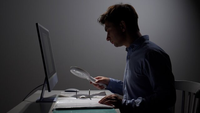 Man examining important legal documents with a magnifying glass while sitting at a desk with a computer in a dimly lit office, focusing on details during an investigation