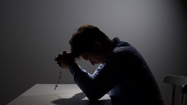 Man holding a rosary and bowing his head in earnest prayer, seeking spiritual guidance and comfort during a moment of intense devotion or personal struggle