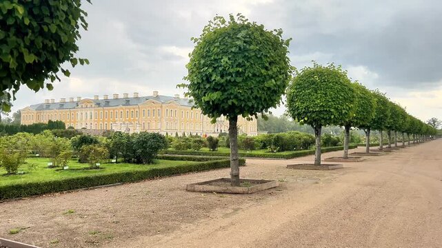 Wide shot of a historic yellow baroque palace seen from a formal garden avenue lined with round pruned topiary trees under a cloudy sky.