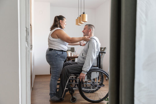 Two individuals engage in dressing assistance at home. Personal assistant offers support to a wheelchair user.