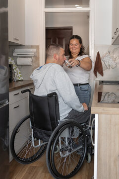 Woman provides personal assistance to man on a wheelchair to drink water in a kitchen. The setting promotes independent living and care.