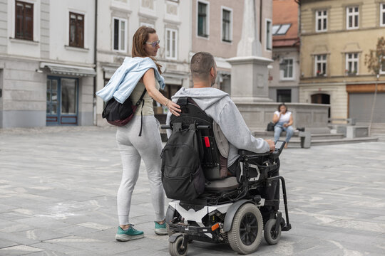 A woman personal assistant supports man in a wheelchair in an urban area. They navigate the square while others sit nearby.