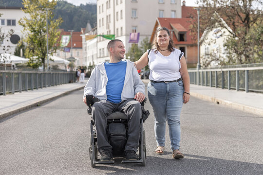 A woman personal assistant assists a man in a wheelchair on a city street during daylight. They are near buildings and walking together.