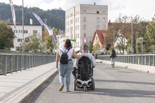 A woman personal assistant walks alongside a man in a wheelchair on a city street with buildings nearby. They navigate the area together.