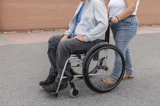 A woman personal assistant helps a man in a wheelchair as they move through an urban area in broad daylight.