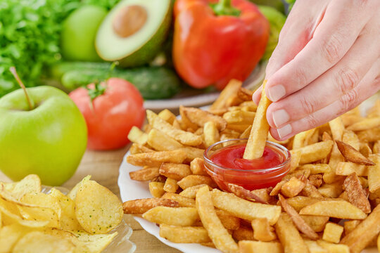 Hand dips a french fry in ketchup beside fresh fruit and vegetables