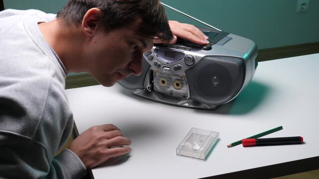 Young man focused on repairing an old cassette boombox player on a white table, carefully inspecting the tape mechanism while pushing buttons to test its functionality