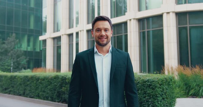 Confident, successful businessman in a formal suit standing outdoors with arms crossed, smiling and looking at the camera in front of a modern glass office building.