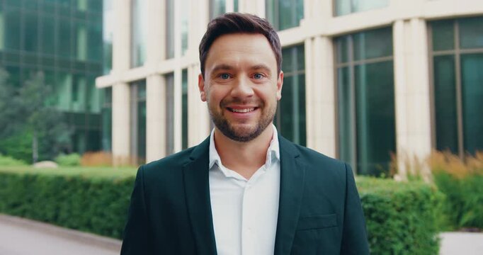 A confident man in formal attire carrying a laptop looks into the camera and smiles while walking through a park near a modern glass building.