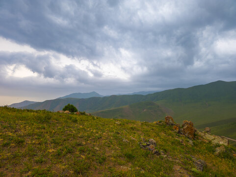 Landscape photograph features rolling green hills under a dramatic overcast sky in Kyrgyzstan. Grassy slopes with rocks create a moody atmosphere, hinting at an impending storm.