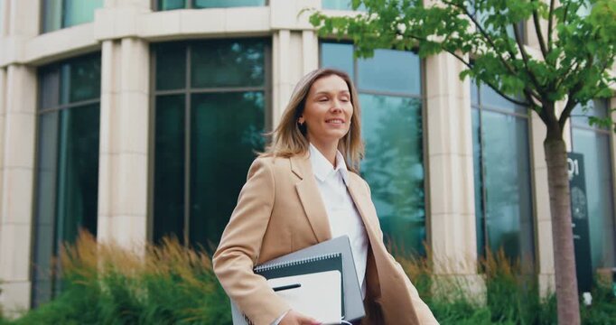 Side view of a young Caucasian woman confidently walking near a modern glass office building, smiling as she carries a laptop and a notebook in her hands. Joyful and determined on her way to work
