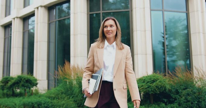 Business woman in a formal suit walking outdoors near a modern office building while using smartphone for online communication. Professional corporate lifestyle, urban business environment, modern