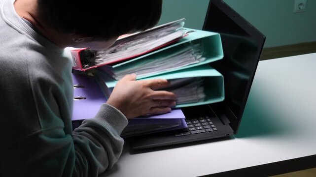 Overwhelmed person expressing frustration and exhaustion, holding a stack of heavy ring binders over a laptop, highlighting the concept of excessive workload and burnout
