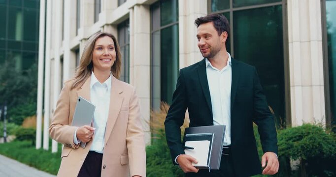 Happy business colleagues, a man and a woman in formal suits, are walking side by side through an urban area, engaged in conversation on their way to work, with the woman carrying a laptop
