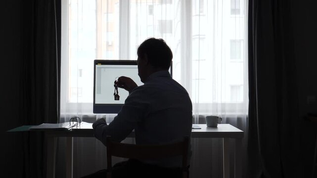 Silhouette of a person sitting at a desk in a home office, holding a house shaped keychain while looking at a computer screen, symbolizing real estate decisions and property ownership