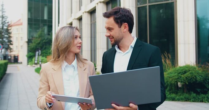 Portrait of two business professionals looking at the camera while standing in a modern startup office, holding a tablet and a laptop.