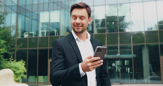 Side view of a young Caucasian man in formal clothes walking on the street while using a tablet, with a modern building in the background.