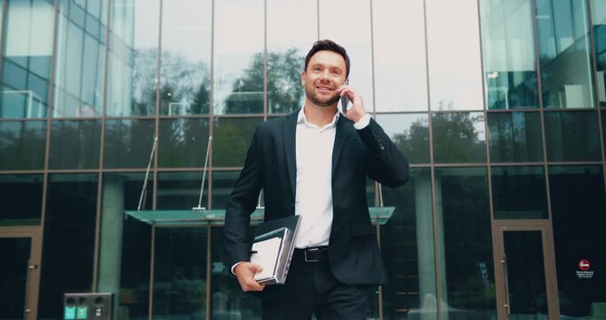 Confident businessman in a formal suit holding a laptop and talking on the phone while walking outdoors in a park area. Modern glass office building in the background. Business communication