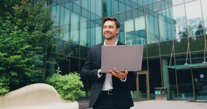 Confident businessman working on a laptop outdoors near a modern glass office building, representing remote work, digital communication, and modern urban business lifestyle