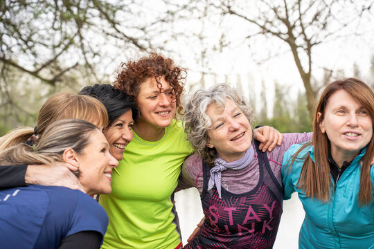 Active generation x and baby boomer women embrace in a cheerful outdoor fitness group. Smiling friends share teamwork, support, friendship, and healthy aging in nature.