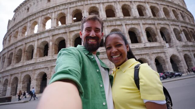 Smiling mixed race couple making a selfie video for social media with colosseum in background, looking at camera cheerfully.