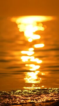 Glistening golden sunset creates vibrant shimmering bokeh reflections on moving sea waves near sandy shoreline. Vertical tripod steady close up shot.