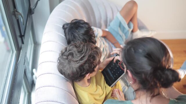 Mother teaching her child using a digital tablet at home, with the other son relaxing on the sofa next to them, representing homeschooling and modern education