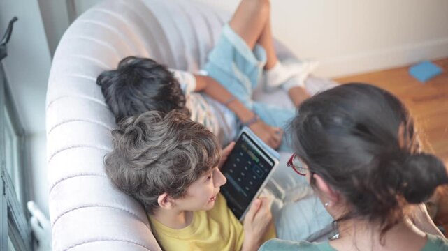 Mother teaching her two young sons how to use a tablet computer at home, family enjoying educational app together, concept of learning and modern technology in childhood