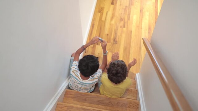 Two boys sitting together on a wooden staircase sharing music and dancing with headphones, experiencing joy and modern technology in their childhood
