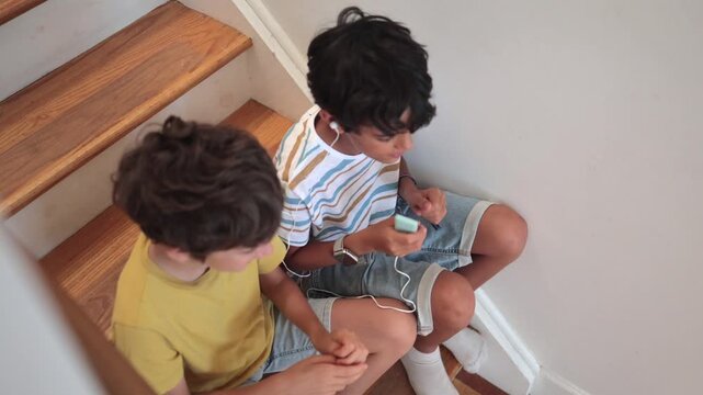 Two young boys sitting on wooden stairs, listening to music on a media player with earphones, joyfully dancing and singing together while enjoying their favorite song at home