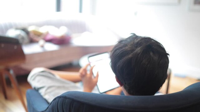 Back view of a boy browsing a tablet, engaged in e-learning while sitting comfortably on a chair at home, with his mother sleeping on the sofa in the background