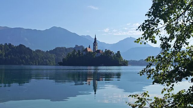 Bled Island and the Church of the Assumption reflected in the calm waters of Lake Bled, Slovenia, with the Julian Alps and trees surrounding the scenic landscape.