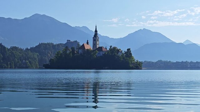 Bled Island and the Church of the Assumption on Lake Bled, Slovenia, surrounded by calm blue water and Alpine mountains, with reflections and sunlight visible on the lake.