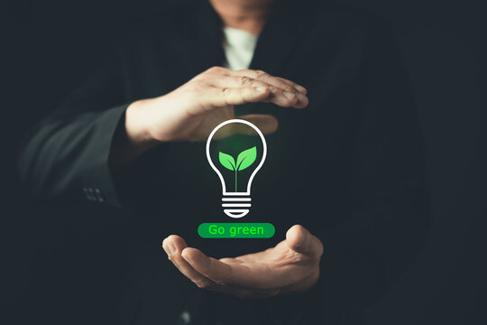 Businessman protecting glowing green lightbulb with plant leaves and go green text on hands, representing renewable clean energy, environmental protection, ESG concept, and sustainable ecology idea.