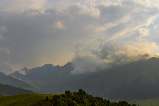 Breathtaking view of a mountain range beneath a dynamic sky filled with clouds. Rolling green hills and evergreen trees lead to imposing, snow-capped peaks in the background.