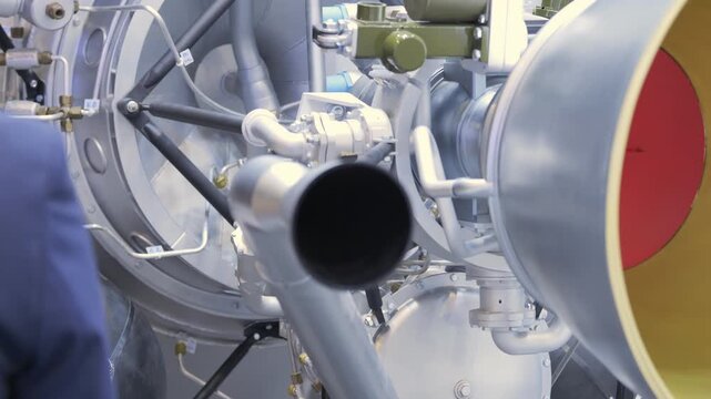 Close-up of mechanical engine parts with pipes and metal components. Red and yellow circular elements visible. Blue uniform partially seen in background. Detailed machinery view